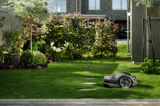 Husqvarna Automower® 305E NERA. Robotic lawn mower operating in a well-maintained garden with a pathway and shrubs.