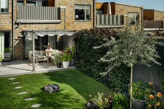 Husqvarna Automower® 305E NERA. A robotic lawn mower operating in a well-maintained garden with a person relaxing on a patio.