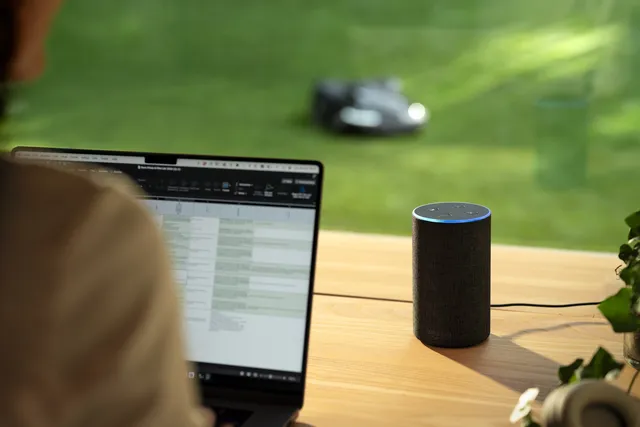 A person working on a laptop with a robotic lawn mower operating in the garden outside.