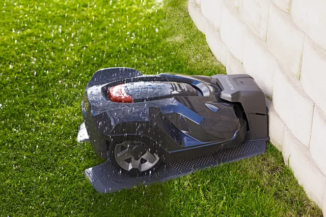 Robotic lawn mower returning to its charging station in a garden, with water droplets on it.
