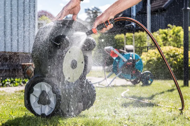 A person cleaning a robotic lawn mower with a hose in a garden setting.